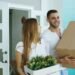 Couple entering their home carrying a cardboard box and indoor plant during move-in