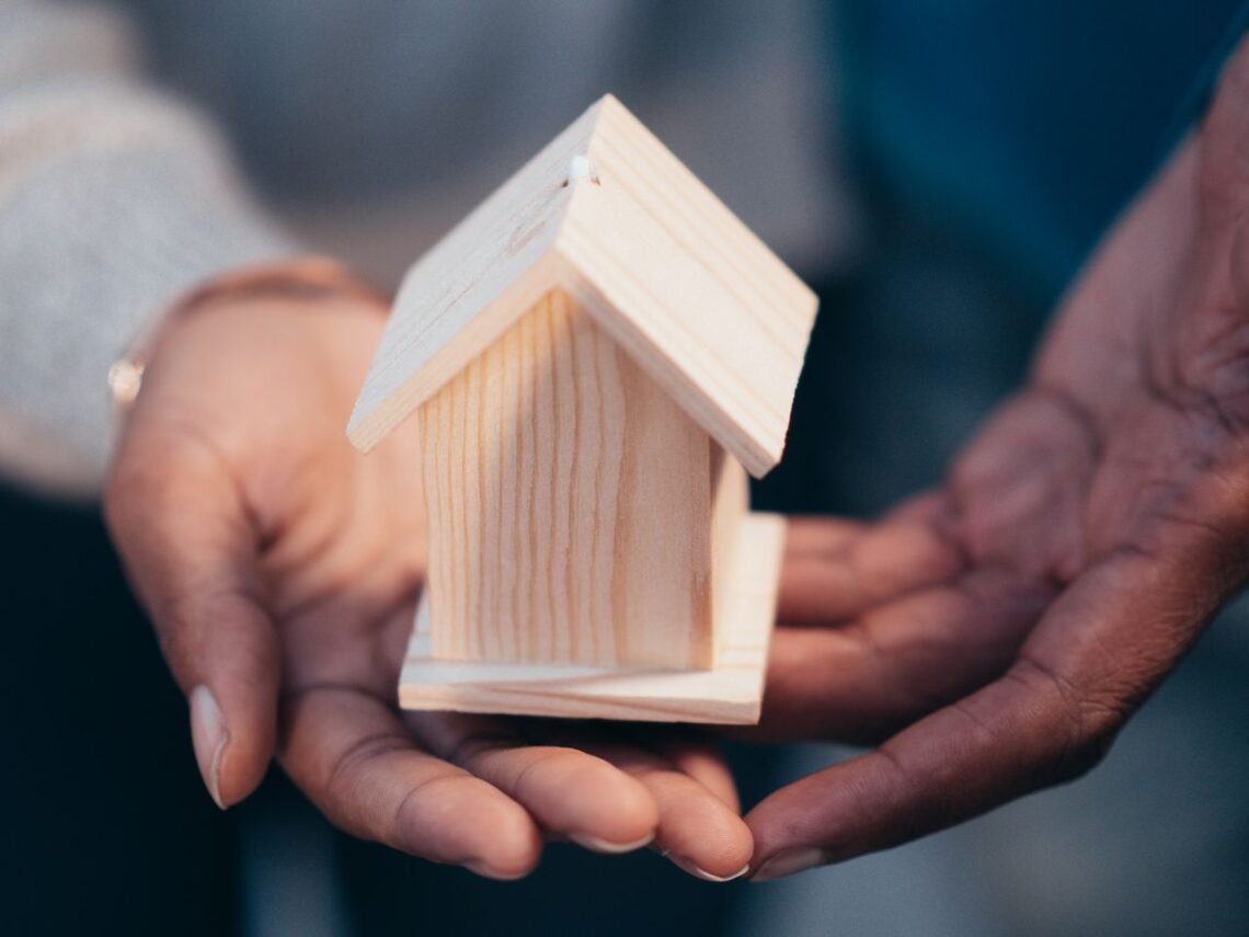 Hands protecting a small wooden house model representing home insurance coverage
