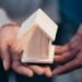 Hands protecting a small wooden house model representing home insurance coverage