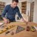 DIY homeowner measuring a wooden board with a tape measure while using essential measuring tools including a spirit level, digital caliper, combination square, laser distance measure, stud finder, and angle finder in a home workshop.